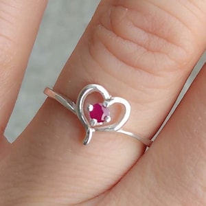 May include: A silver ring with a heart-shaped design and a small red gemstone in the center.