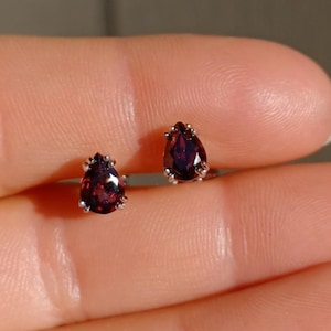 May include: A pair of stud earrings featuring deep red, pear-shaped garnet gemstones set in silver-toned metal prongs. Each teardrop-shaped stone is faceted to reflect light, showcasing a rich, dark crimson hue held securely by small metallic claws.