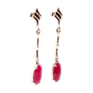 Op de afbeelding: Een paar zilverkleurige oorhangers. Elke oorbel heeft een decoratief bovenstuk, een bamboe-achtige steel en een levendige, ovaalvormige, rode edelsteen aan de onderkant. De oorbellen worden tegen een witte achtergrond getoond.