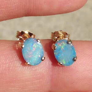 May include: A pair of gold stud earrings featuring oval-shaped opals. The opals have a light blue base color with iridescent flashes of green, pink, and yellow. The earrings are set in a gold metal.