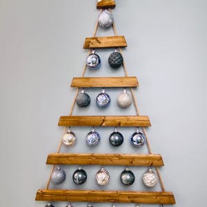 May include: A wooden Christmas tree-shaped wall hanging with a star topper. The tree is constructed of stained wooden planks and rope, holding various silver, gray, and white ornaments. The tree is mounted on a light gray wall.