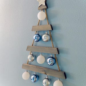 以下が含まれることがあります： 木製のクリスマスツリーの装飾。上部に星があり、灰色の木の板とロープで作られ、白、銀、青のオーナメントで飾られています。明るい青色の壁に掛けられています。