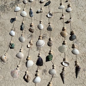 Könnte beinhalten: Dekorative Wanddekoration mit einer Bambusstange, Schnüren, Muscheln und Holzperlen. Die Muscheln sind in verschiedenen Formen, Größen und Farben, darunter Weiß, Beige und Grau. Für die Inneneinrichtung.