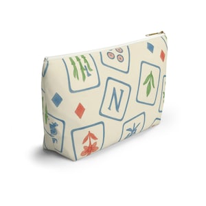 Puede incluir: Bolsa con cremallera de color crema con un patrón repetido de cuadrados enmarcados en azul que contienen varias ilustraciones de acuarela. Las ilustraciones incluyen bambú, flechas y la letra "N", junto con rombos rojos y azules.