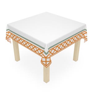 Puede incluir: Un mantel cuadrado blanco con un diseño de borde geométrico naranja y verde. El mantel está colocado sobre una mesa cuadrada con patas de madera de color claro. El diseño presenta un patrón de diamantes repetido.