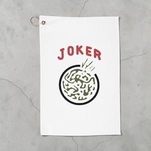 以下が含まれることがあります： 白いゴルフ用タオル。赤い「JOKER」の文字が、緑と黒のデザインが入った黒い円の上に配置されています。吊り下げ用の金属製グロメット付き。