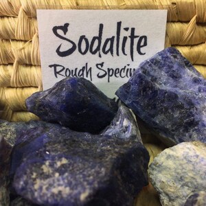 May include: A collection of rough sodalite crystals in various shades of blue. The crystals are arranged in a basket and have a natural, unpolished appearance. The label reads "Sodalite Rough Specimen".
