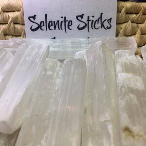 May include: A close-up of a group of white selenite sticks. The sticks are smooth and have a natural, striated texture. The sticks are arranged in a basket, and the text "Selenite Sticks" is visible on a white card.