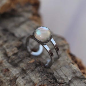 May include: A silver ring with a round, iridescent, opalescent stone. The ring has a double-band design and is set against a textured, brown background.