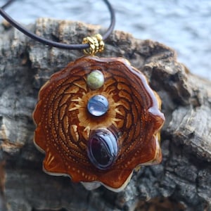 May include: A handcrafted pendant necklace featuring a brown, carved wooden pendant with three embedded gemstones: green, blue, and dark red. The pendant is suspended from a dark brown leather cord with a gold-colored clasp.