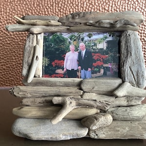 May include: A rustic photo frame crafted from weathered driftwood. The frame surrounds a photograph of a couple in a garden setting. The frame's design incorporates various sizes and shapes of driftwood pieces, creating a natural, textured appearance. The background is a textured copper surface.