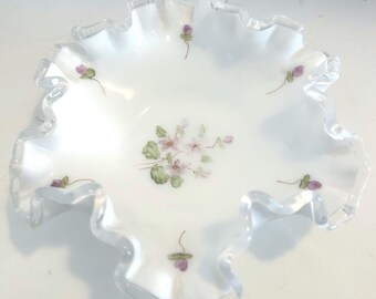 Puede incluir: Un cuenco de vidrio blanco con borde ondulado y diseño floral. El cuenco presenta un delicado patrón de pequeñas flores moradas y hojas verdes. El borde tiene un borde ondulado y festoneado.