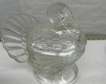 May include: Clear glass turkey-shaped dish with a textured body and tail feathers. The dish sits on a round base and features a detailed head and neck. A decorative piece for serving or display.
