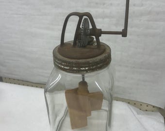Könnte beinhalten: Ein alter Butterfass aus Glas mit Metallmechanismus und Holzgriff. Das Fass hat ein quadratisches Glasgefäß mit einem Metalldeckel und einem Holzpaddel im Inneren. Die Metallkomponenten weisen Gebrauchsspuren auf.