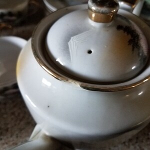 May include: White teapot with a gold trim and a lid with a floral design. The teapot has a spout and a handle.