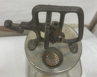 Könnte beinhalten: Ein antikes Passiergerät aus Gusseisen mit einem Glasbehälter. Es hat eine Kurbel, Zahnräder und eine perforierte Metallplatte. Die Metallteile weisen eine verwitterte, Vintage-Optik auf.