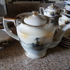 May include: A white teapot with a gold trim and a Japanese landscape design. The teapot is sitting on a granite countertop with other teacups and a sugar bowl.