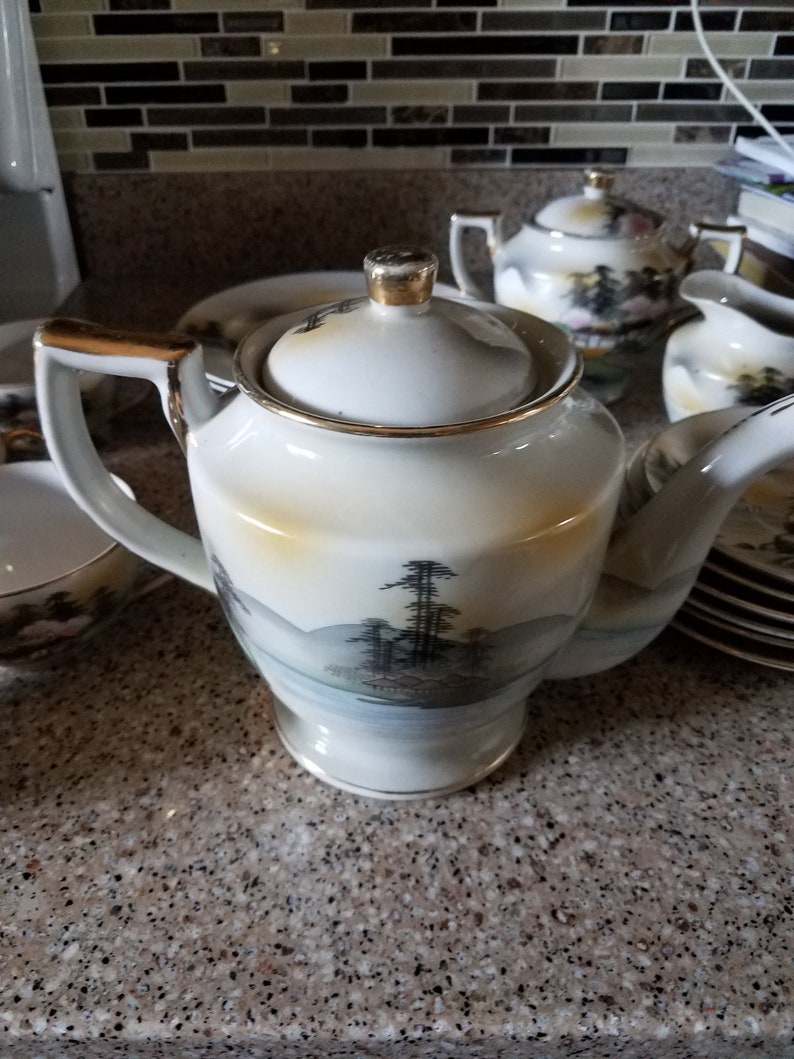 May include: A white teapot with a gold trim and a Japanese landscape design. The teapot is sitting on a granite countertop with other teacups and a sugar bowl.