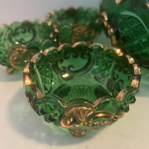 Puede incluir: Juego de cuatro saleros de vidrio verde con ribetes dorados y borde festoneado. Los saleros tienen un diseño decorativo y se apoyan en tres pequeñas patas.