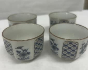 Könnte beinhalten: Vier kleine, weiße Keramikbecher mit blauem Blumendesign und geometrischem Muster. Die Becher haben einen goldenen Rand und sind quadratisch angeordnet. Die Becher sind wahrscheinlich für Tee oder Sake.
