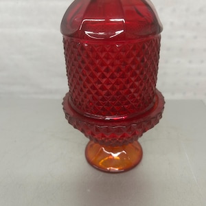 Puede incluir: Un portavelas de cristal rojo vibrante con un patrón de diamantes texturizado en el cuerpo cilíndrico y una base acampanada. La sección superior tiene un borde liso y redondeado, mientras que la parte central muestra un diseño geométrico.