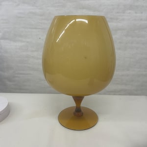 May include: A large, amber-colored glass goblet with a rounded bowl and a stem. The goblet sits on a circular base. The glass has a smooth, glossy finish. The overall shape is reminiscent of a brandy snifter.