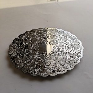 Op de afbeelding: Een zilverkleurige metalen onderzetter met een ingewikkeld bloemmotief. De onderzetter heeft een gegolfde rand en is perfect om oppervlakken te beschermen tegen hitte.
