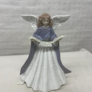 May include: A ceramic angel figurine with brown hair, white wings, and a flowing white dress. The angel wears a periwinkle blue shawl and holds a scroll. The figurine has a detailed design and is likely a decorative item.