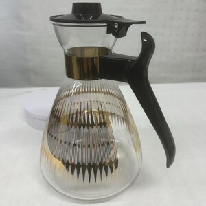 May include: A vintage glass coffee carafe with a black handle, lid, and gold accents. The clear glass body features a gold and white geometric design. The carafe is designed for pouring and serving.