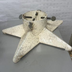Könnte beinhalten: Ein sternförmiger Weihnachtsbaumständer mit cremefarbener Oberfläche und dunkelbraunen Sprenkeln. Die zylindrische Halterung in der Mitte verfügt über vier verstellbare Metallschrauben mit flachen, flügelförmigen Köpfen zur Befestigung.