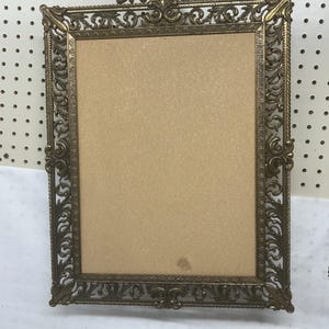 Puede incluir: Un marco de fotos rectangular de metal en tono latón con un borde calado intrincado con volutas y una cresta en forma de concha en la parte superior. El marco contiene una inserción rectangular de color tostado claro con una mancha.