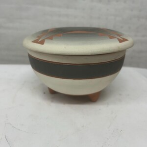Può includere: Una ciotola in ceramica con coperchio, con base color crema e una fascia grigio scuro. Il coperchio è color crema con un motivo geometrico arancione e grigio. La ciotola poggia su tre piccoli piedini arrotondati.