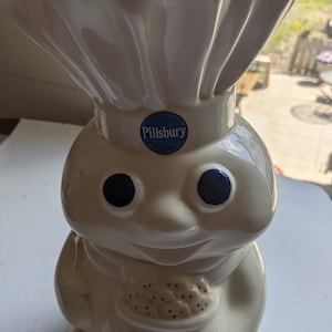 Könnte beinhalten: Weiße Keramik-Keksdose in Form des Pillsbury Doughboy, der einen Teller mit Keksen hält. Die Dose hat blaue Augen, ein blaues Pillsbury-Logo und das Wort "COOKIES" in Blau auf der Vorderseite.