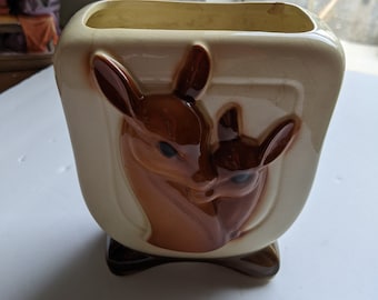 Może przedstawiać: Ceramiczna doniczka z brązową i białą glazurą przedstawia reliefowy wzór dwóch jeleni, jeden większy od drugiego. Jelenie patrzą na widza i mają brązowe oczy i nosy.