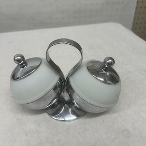 Peut inclure: Un ensemble de salière et poivrière vintage. L'ensemble comprend deux récipients en verre sphériques blancs avec des bords en métal argenté et un petit bouton sur le dessus. Les récipients sont reliés par une poignée et une base argentées.