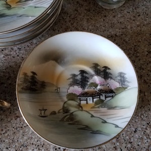 May include: A white porcelain saucer with a gold rim features a hand-painted scene of a Japanese village with houses, trees, and a river. The scene is painted in shades of green, brown, and pink.