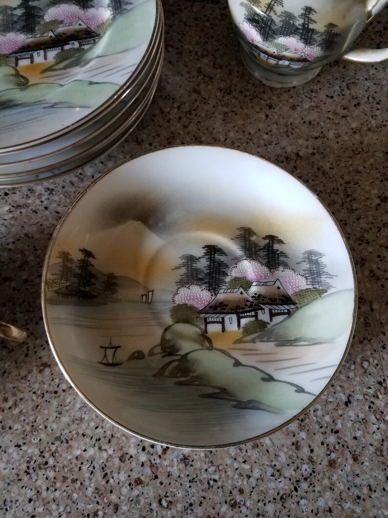 May include: A white porcelain saucer with a gold rim features a hand-painted scene of a Japanese village with houses, trees, and a river. The scene is painted in shades of green, brown, and pink.