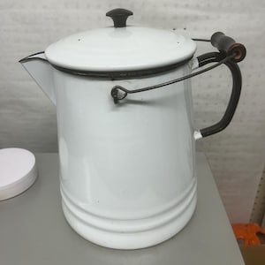May include: A white enamel coffee percolator with a black rim, a black lid knob, and a black metal handle. The cylindrical body features horizontal ridges near the base and a spout on the left side. The metal handle is attached with a wire frame.