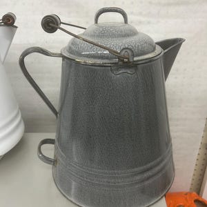 以下が含まれることがあります： 斑点模様の仕上げが施されたグレーのエナメル製コーヒーポット。先細りの本体、サイドハンドル、注ぎ口、そして上部のワイヤーフレームに2つの小さな木製ノブが付いたワイヤーベイルハンドル付きの金属製蓋が特徴です。
