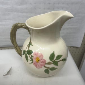Peut inclure: Pichet en céramique crème avec un motif floral. Le pichet présente une rose rose avec un centre jaune et des feuilles vertes. La poignée est conçue pour ressembler à une branche d'arbre.