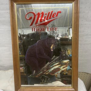 Puede incluir: Un espejo enmarcado con un marco de madera marrón. El espejo presenta el logotipo de Miller High Life en escritura roja. Debajo, una representación de un oso negro sobre un tronco, con un oso más pequeño en el fondo. Las palabras "Black Bear Wisconsin" están en la parte inferior.