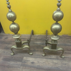 May include: Pair of antique brass fireplace andirons. Each andiron features a decorative, tiered finial atop two spherical elements. The base has curved legs and a rectangular support. The andirons are displayed on a wood-grain surface against a yellow wall.