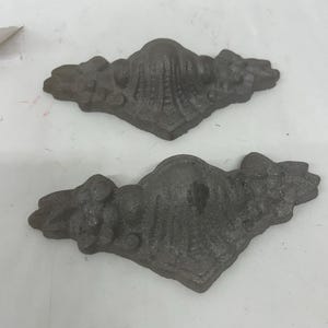 Può includere: Due staffe angolari decorative in ghisa di colore grigio scuro. Le staffe presentano una superficie strutturata e un motivo floreale. Sono simmetriche e di forma curva.