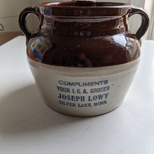 May include: A brown and white ceramic crock with a lid. The crock has blue lettering that reads "Compliments Your I.G.A. Grocer Joseph Lowy Silver Lake, Minn."