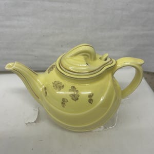 Puede incluir: Una tetera de cerámica amarilla con asa y boquilla curvadas. La tetera tiene tapa y está decorada con diseños de hojas doradas. La tetera es de color amarillo claro.