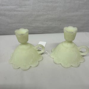 Puede incluir: Dos candelabros de vidrio amarillo pálido. Cada uno tiene una base festoneada con un patrón decorativo y un pequeño asa. La parte superior de cada soporte tiene una copa en forma de tulipán. Los candelabros son de vidrio esmerilado.