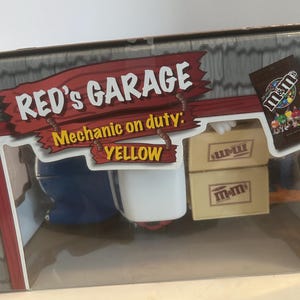 Puede incluir: Una caja de cartón con un letrero rojo y amarillo que dice "RED'S GARAGE Mecánico en servicio: AMARILLO". La caja contiene un coche de juguete y dos cajas de caramelos M&M's.
