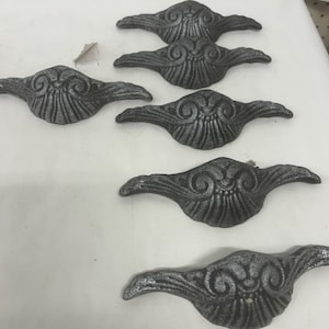 Può includere: Sei pomelli decorativi per cassetti in metallo pressofuso con finitura argentata. Ogni pomello presenta un design simmetrico con un ricciolo centrale e ali stilizzate. Sono disposti su una superficie bianca, per mobili.