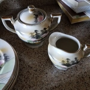 May include: A set of white porcelain teacups and saucers with a delicate floral and landscape design. The set includes a sugar bowl with a lid and a creamer.