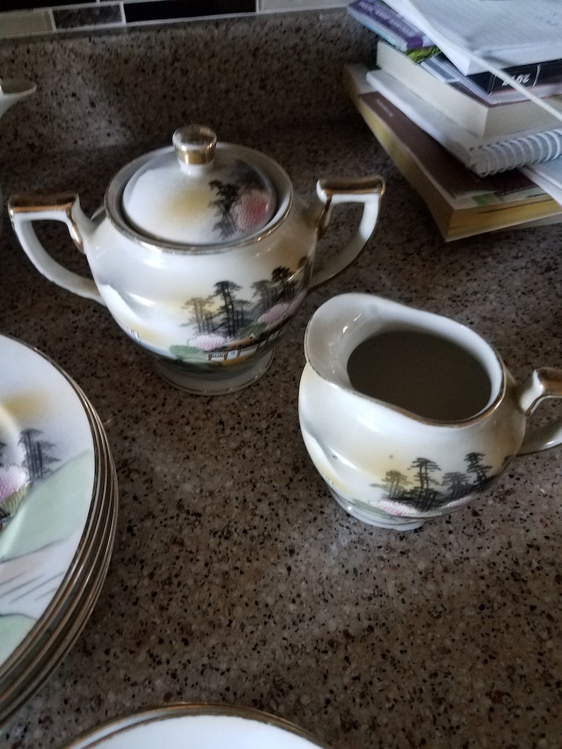 May include: A set of white porcelain teacups and saucers with a delicate floral and landscape design. The set includes a sugar bowl with a lid and a creamer.